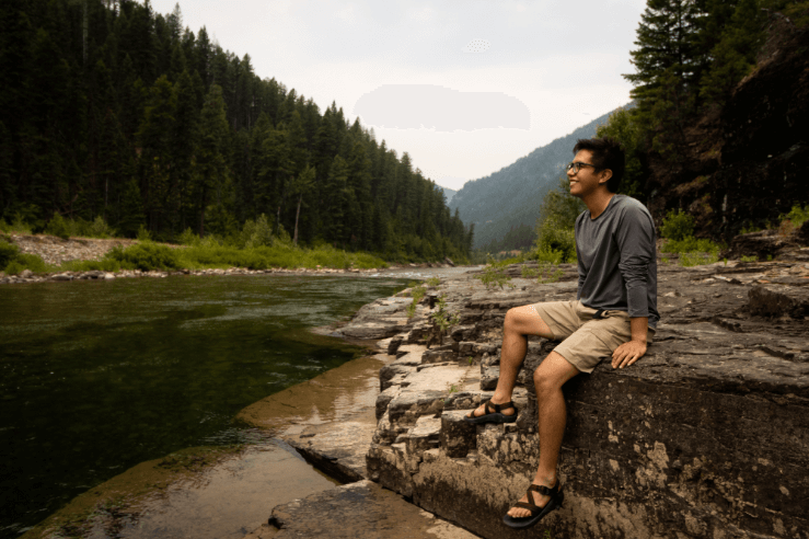 A man sitting on a rocky bank by a river surrounded by a lush green forest