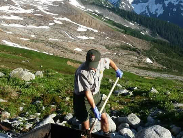 National Park Service employee working in a mountain wilderness with snow-covered peaks in the background.