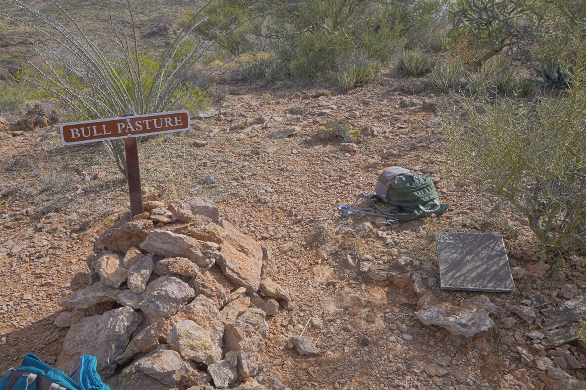 A metal sign sticking out of a large pile of rocks with a backpack on the trail