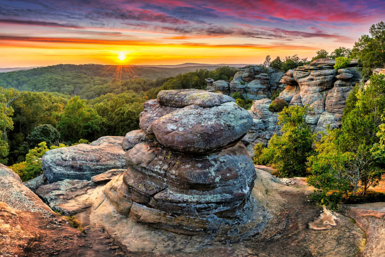 Large weathered rock outcroppings in the foreground with a rolling green forest in the background under a beautiful sunrise